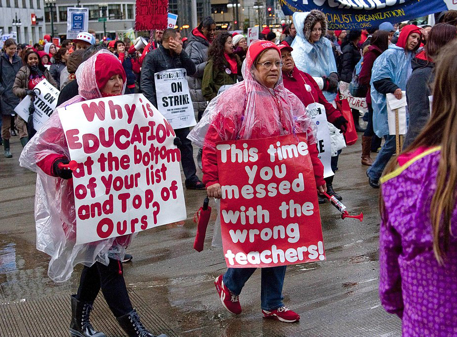 Des enseignants en vêtements de pluie manifestant dans la rue avec des banderoles et des panneaux de piquetage.  Un personnage au premier plan porte une pancarte disant : « Pourquoi l'éducation est-elle en bas de votre liste et en haut de la nôtre ?  Le signe de la deuxième figure au premier plan se lit comme suit : 