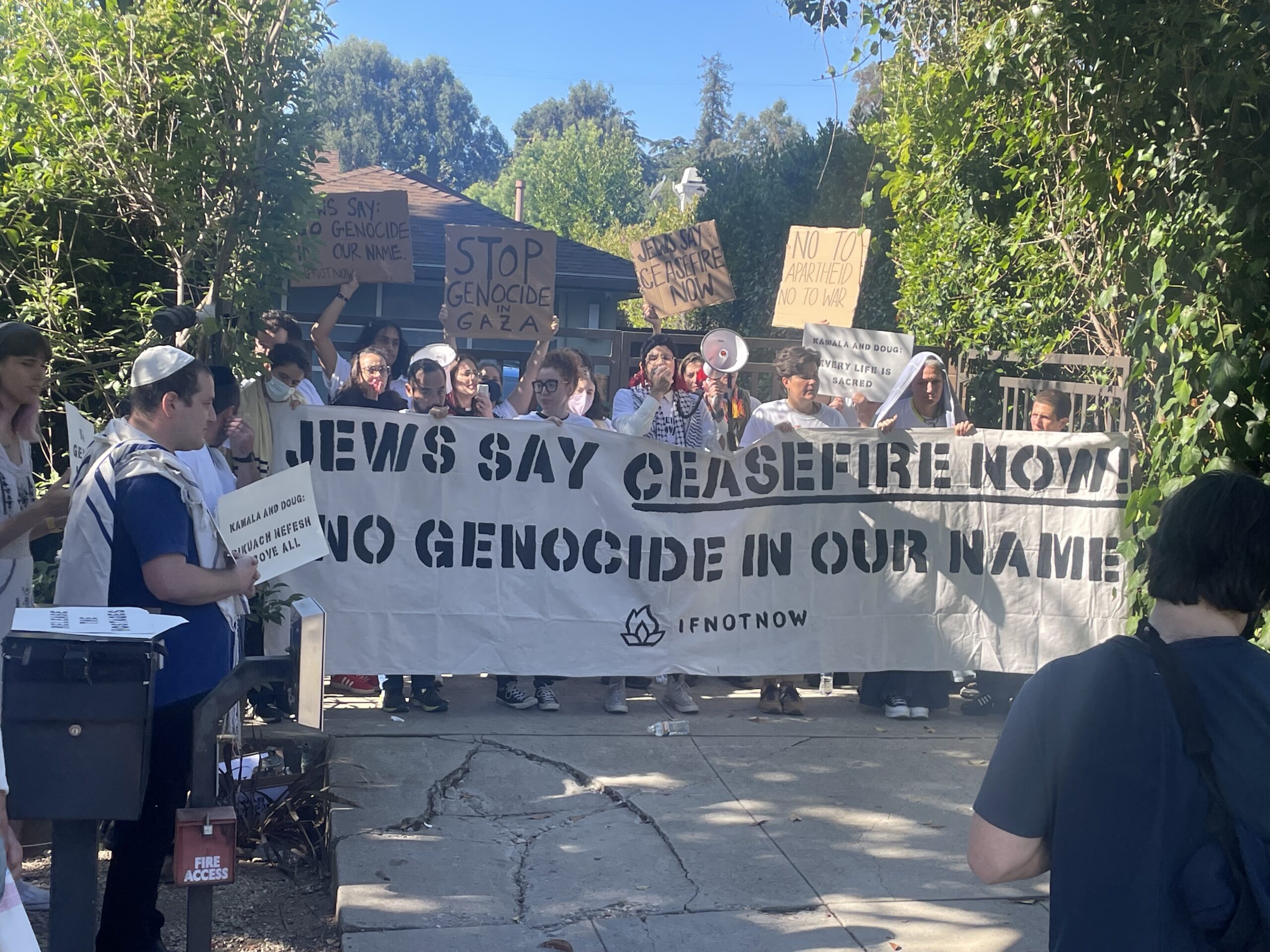 Une cinquantaine de personnes brandissant des pancartes indiquant Stop au génocide à Gaza se tiennent derrière une banderole en noir et blanc sur laquelle on peut lire Les Juifs disent cessez-le-feu maintenant.  Pas de génocide en notre nom.  SI PAS MAINTENANT